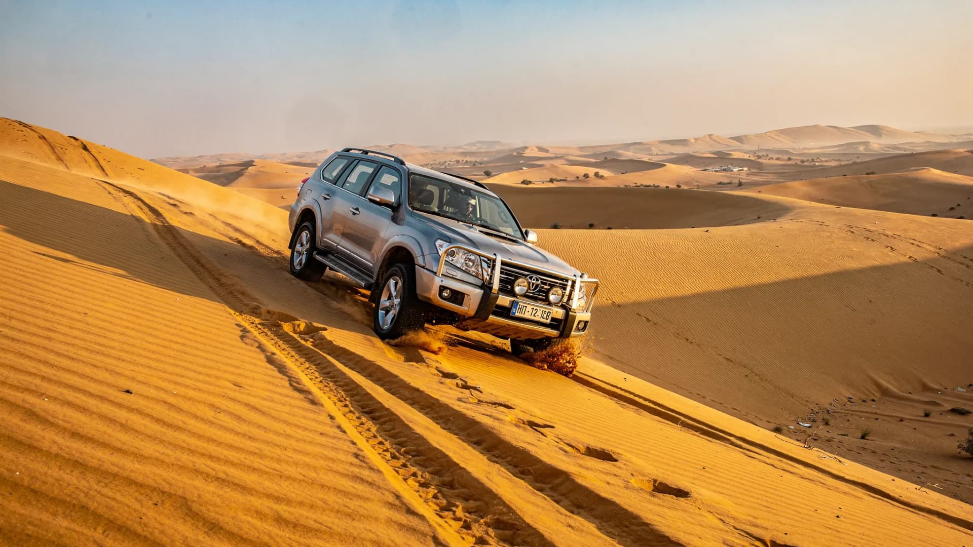 A 4x4 SUV descending a steep sand dune in the Dubai desert, with clear tire tracks behind it and rolling dunes stretching into the distance.