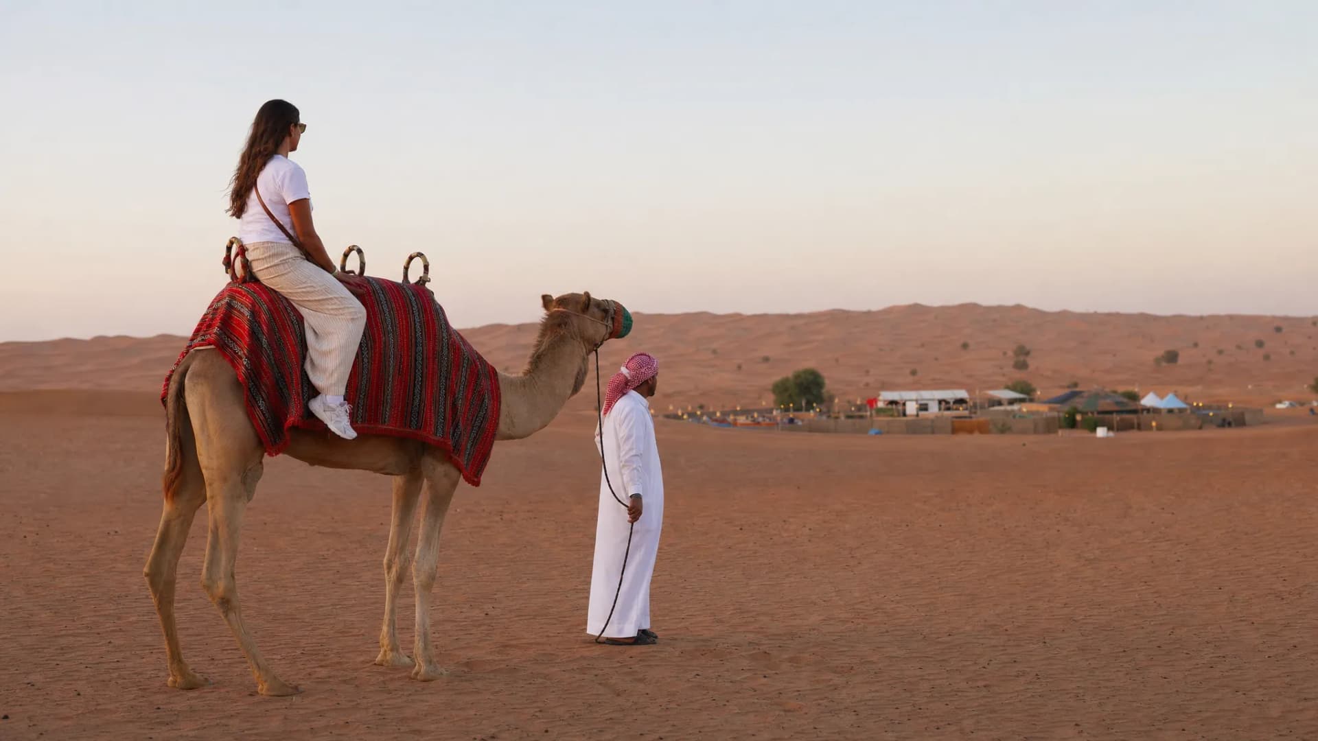 A single camel with a rider and guide paused on the red dunes of Lahbab Desert, with the camel facing sideways and the desert camp far in the background.