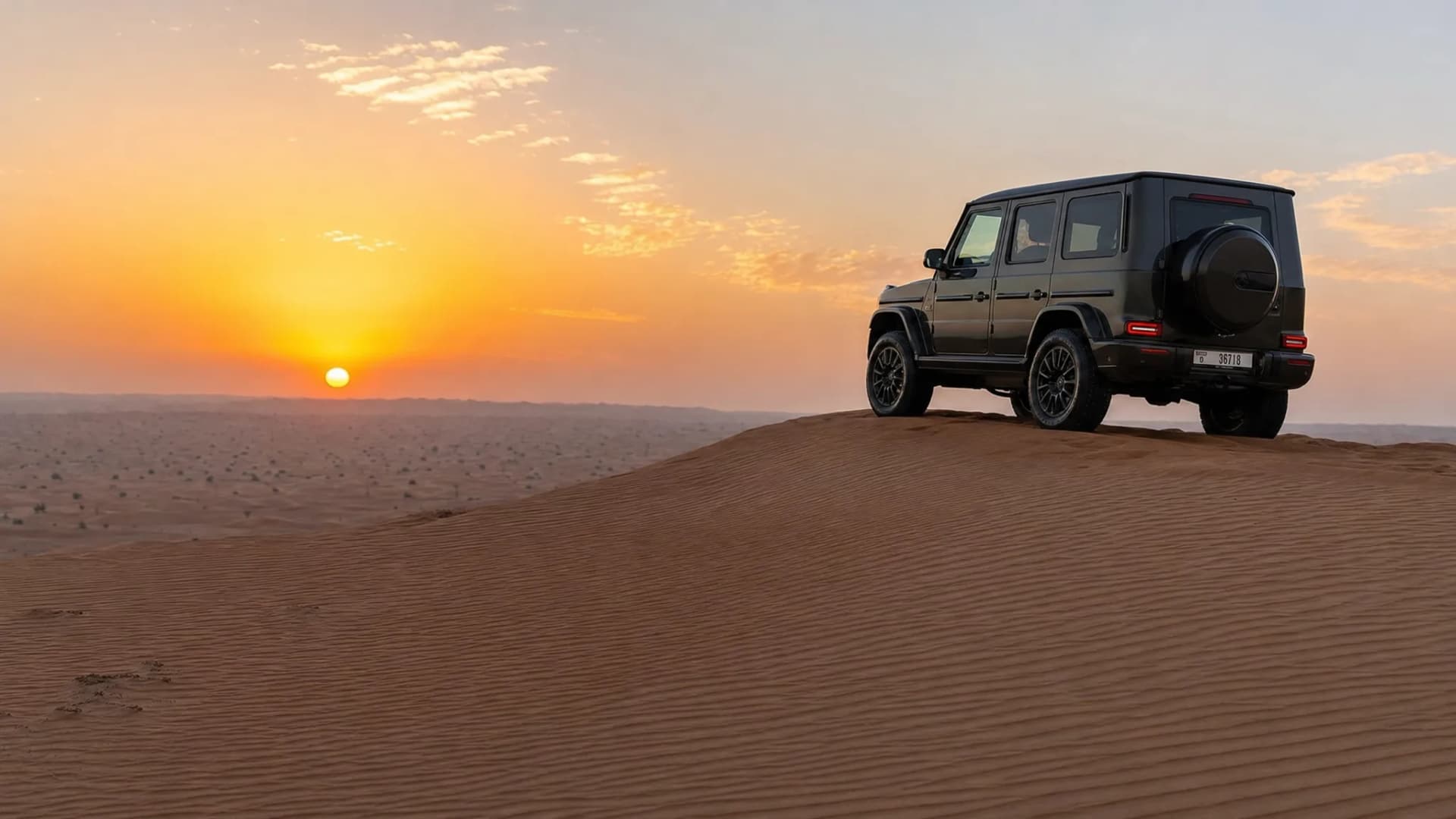 A luxury 4x4 vehicle parked on a high dune in the Lahbab Desert at sunset, with layered red sand patterns and a wide desert horizon in the background.