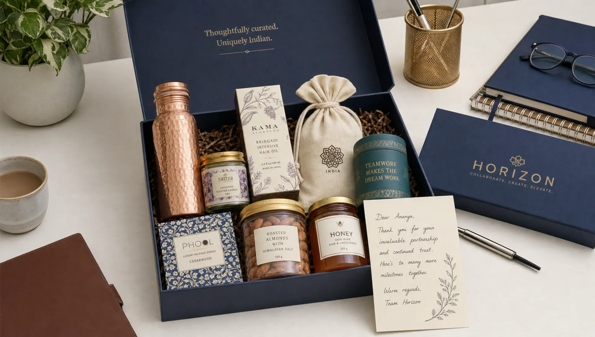 A premium corporate gift box arranged with Indian artisan products, wellness items, gourmet treats, a handwritten note card, and elegant branded packaging on a clean desk.